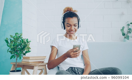 Happy African American woman is surfing internet with smartphone and laughing sitting on comfortable sofa at home, girl is listening to music through headphones. Happy African American woman is surfing internet with smartphone and laughing sitting on comfortable sofa at home, girl is listening to music through headphones. 96099104