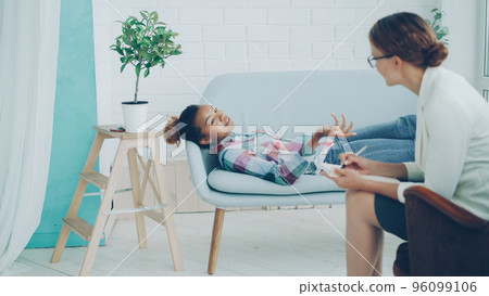 Relaxed African American teenager is talking to psychologist lying on couch and smiling while therapist is listening and taking notes. Young people, mental health and profession concept. Relaxed African American teenager is talking to psychologist lying on couch and smiling while therapist is listening and taking notes. Young people, mental health and profession concept. 96099106
