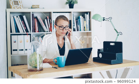 Confident young woman company employee is talking on mobile phone, using laptop and taking notes in notebook sitting at desk at workplace. Business, modern office and people concept. Confident young woman company employee is talking on mobile phone, using laptop and taking notes in notebook sitting at desk at workplace. Business, modern office and people concept. 96099124
