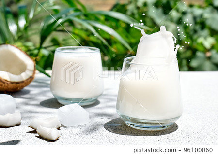 Coconut milk splash in drinking glass on palm leaf background. Vegetable milk, lactose free non dairy healthy drink. Vegan food. 96100060
