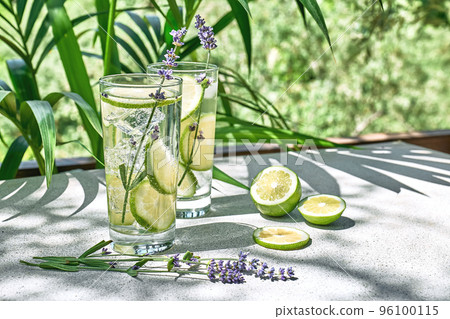 Cool lavender lemonade with lime slices and lavender flower on the table in tropical garden with palm. Healthy organic summer soda drink. Detox water. Diet unalcolic coctail. 96100115