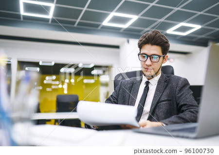 Young office worker in glasses reads documents in the office. Stylish man analyze documents, plans. Young office worker in glasses reads documents in the office. Stylish man analyze documents, plans. 96100730