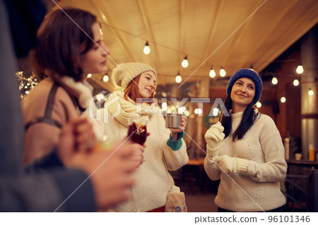 Group of young people spending time together, drinking mulled wine, talking, laughing at winter fair at evening time. Winter holidays, Christmas vibes concept 96101346