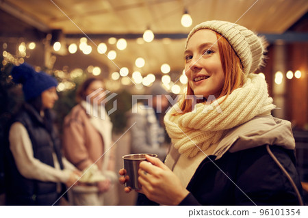 Close-up of young beautiful girl in winter attire drinking hot drinks and dreaming. Spending time with friends at winter fair at evening. 96101354
