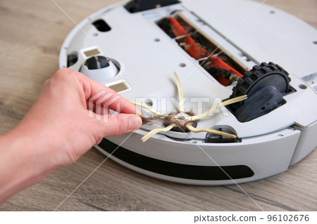 A woman  removes hair from the brush of a robot vacuum cleaner. The concept of a robot vacuum cleaner cleans the floors in the house. 96102676