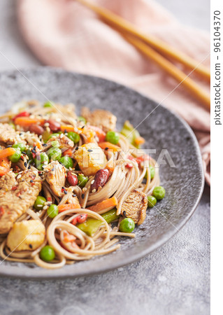 Wok with turkey meat, soba noodles, corn, green peas, green beans and carrots served on gray background with chopsticks. Asian food, concept of street food 96104370