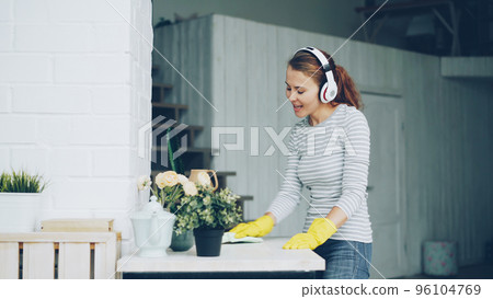 Good-looking girl is dusting table at home and listening to pop music through headphones, singing and dancing with happy face. Millennials, household and interior concept. 96104769