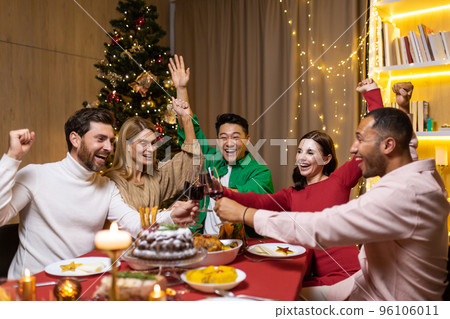 Young cheerful group of interracial people celebrating the coming new year. They sit at the festive table, clink glasses, raise their hands, congratulate each other, laugh, rejoice. Young cheerful group of interracial people celebrating the coming new year. They sit at the festive table, clink glasses, raise their hands, congratulate each other, laugh, rejoice. 96106011