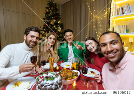 Festive selfie of a group of interracial people. Young men and women are sitting at the table in the apartment near the Christmas tree, holding glasses, looking at the camera, smiling. Festive selfie of a group of interracial people. Young men and women are sitting at the table in the apartment near the Christmas tree, holding glasses, looking at the camera, smiling. 96106012