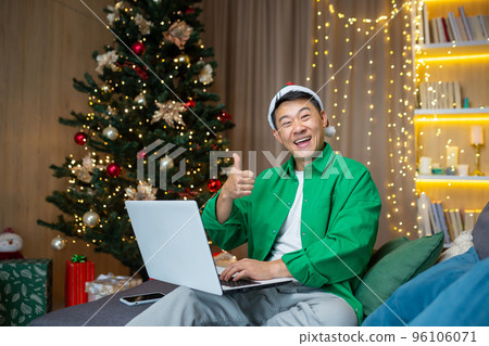 Portrait of a young handsome Asian man sitting on the sofa at home near the Christmas tree in a red hat, holding a laptop and a phone, looking at the camera, smiling, pointing super. Portrait of a young handsome Asian man sitting on the sofa at home near the Christmas tree in a red hat, holding a laptop and a phone, looking at the camera, smiling, pointing super. 96106071