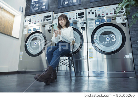 Young woman sitting and looking at smartphone while waiting for laundry to finish at laundromat 96106260
