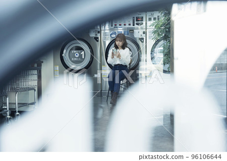 Young woman sitting and looking at smartphone while waiting for laundry to finish at laundromat Young woman sitting and looking at smartphone while waiting for laundry to finish at laundromat 96106644