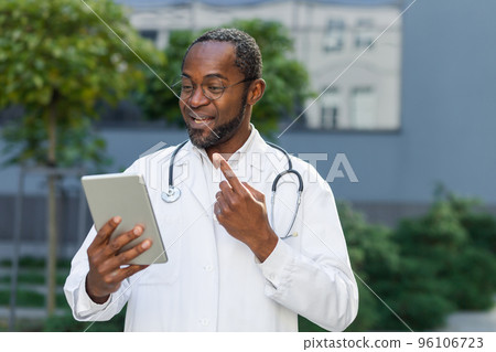 Smiling senior mature doctor in medical coat and glasses with tablet talking on video call remotely online, african american man outside clinic consulting patients. Smiling senior mature doctor in medical coat and glasses with tablet talking on video call remotely online, african american man outside clinic consulting patients. 96106723