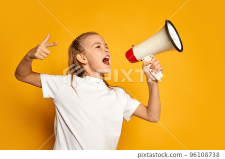 Portrait of little beautiful girl, child in white T-shirt posing, loudly shouting in megaphone isolated over yellow studio background Portrait of little beautiful girl, child in white T-shirt posing, loudly shouting in megaphone isolated over yellow studio background 96108738