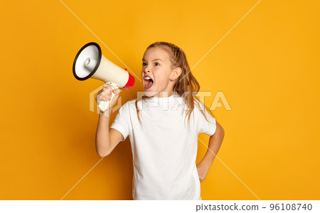 Portrait of little beautiful girl, child in white T-shirt posing, shouting in megaphone isolated over yellow studio background 96108740