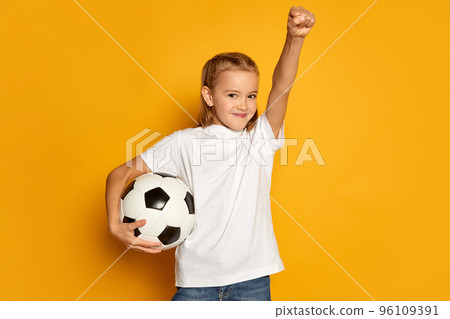 Portrait of little beautiful girl, child in white T-shirt posing with football ball isolated over yellow studio background. Winning 96109391