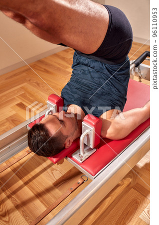 Fit man doing a lunge stretch yoga pilates exercise to strengthen and tone his muscles using a reformer in a gym Fit man doing a lunge stretch yoga pilates exercise to strengthen and tone his muscles using a reformer in a gym 96110953