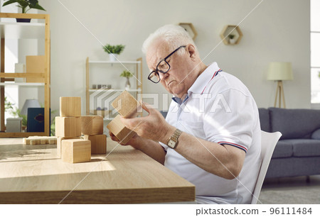 Demented senior man with Alzheimer's disease sitting at table and playing with wooden cubes Demented senior man with Alzheimer's disease sitting at table and playing with wooden cubes 96111484