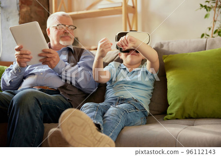 Portrait of happy senior man and little boy, grandfather and grandson using VR glasses sitting on sofa at home and laughing. Hobbies, leisure activities, family 96112143