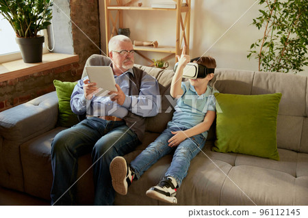 Old man and little boy, grandfather and grandson in casual clothes sitting on sofa at home and using VR glasses. Concept of hobbies, leisure activities, family Old man and little boy, grandfather and grandson in casual clothes sitting on sofa at home and using VR glasses. Concept of hobbies, leisure activities, family 96112145