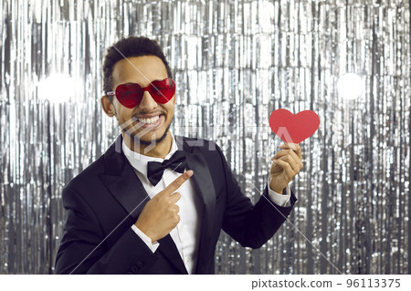 Attractive man in classic suit shows red heart in his hand standing on shiny silver background. Attractive man in classic suit shows red heart in his hand standing on shiny silver background. 96113375