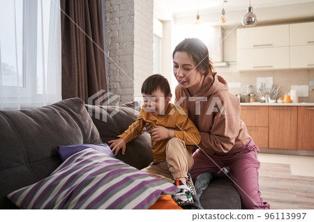 Mother carefully helping to her lovely son while they sitting at the sofa Mother carefully helping to her lovely son while they sitting at the sofa 96113997