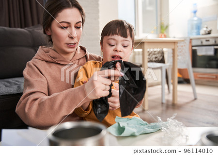 Kid with down syndrome sorting garbage near his mother at the table at home 96114010
