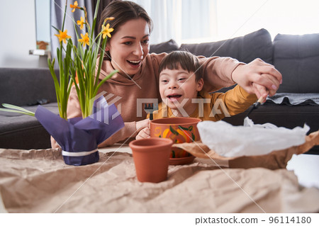 Little boy with down syndrome planting flowers in a brown flowerpots with his mother 96114180