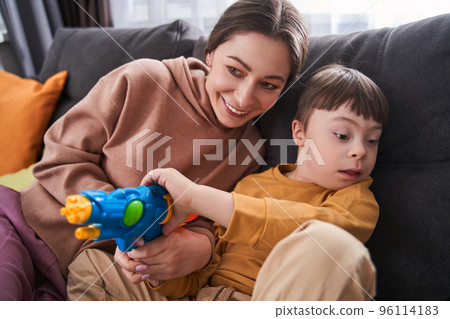 Little boy with down syndrome sitting at the sofa near his cheerful mother and playing 96114183