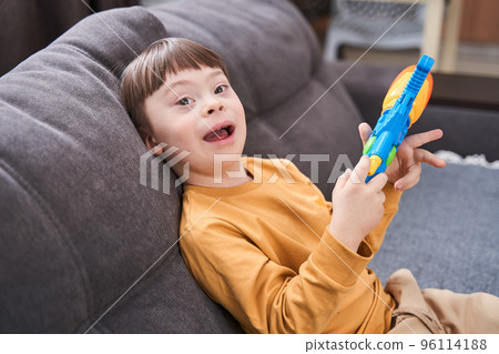 Boy with genetic disorder sitting at the sofa and laughing while playing with toy gun 96114188