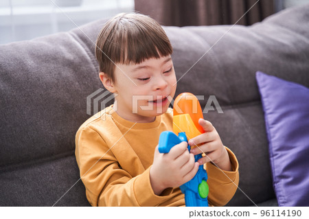 Kid with down syndrome sitting at the sofa and looking at his toy gun 96114190
