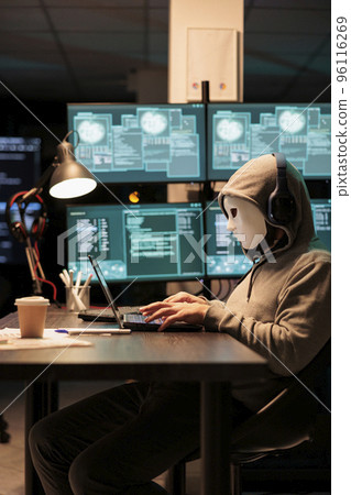 Dangerous masked hacker stealing information from database server online, hacking computer hardware system. Impostor with hood and mask creating malware and security breach in office. 96116269