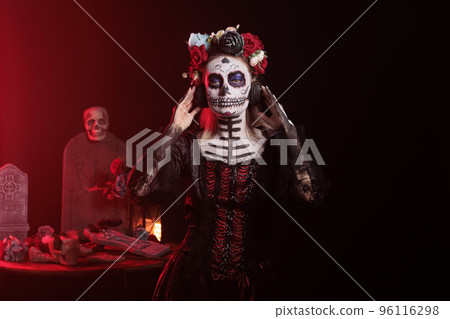 Goddess of death enjoying music on headphones, wearing skull make up and santa muerte costume in studio. Creepy woman listening to song on audio headset, celebrating day of the dead. Goddess of death enjoying music on headphones, wearing skull make up and santa muerte costume in studio. Creepy woman listening to song on audio headset, celebrating day of the dead. 96116298