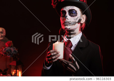 Person in santa muerte lady costume blowing candle flame, posing for dios de los muertos, medican holy celebration in studio. Looking scary and horror with fire, traditional body art paint. 96116303