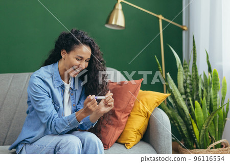 Happy woman alone at home happy with pregnancy test result, Hispanic woman alone at home sitting on couch in living room. Happy woman alone at home happy with pregnancy test result, Hispanic woman alone at home sitting on couch in living room. 96116554