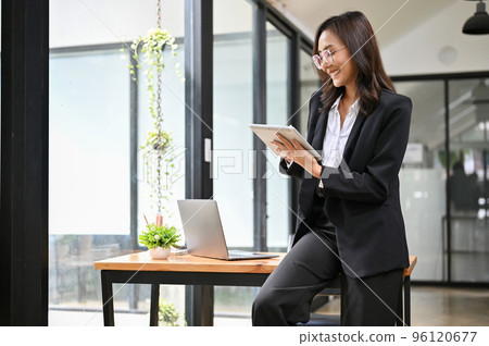 Professional Asian businesswoman in formal suit sitting on her desk, using her tablet. Professional Asian businesswoman in formal suit sitting on her desk, using her tablet. 96120677
