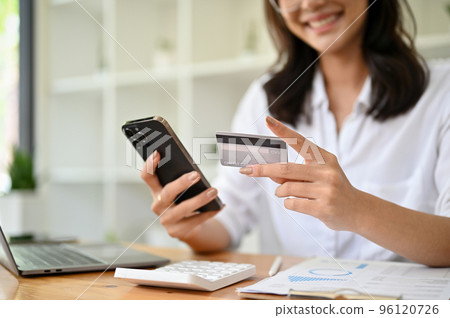 Happy Asian businesswoman at her office desk holding a mobile phone and a credit card Happy Asian businesswoman at her office desk holding a mobile phone and a credit card 96120726