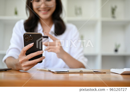 Attractive Asian female sits at her desk, holding a smartphone and a credit card. Online payment Attractive Asian female sits at her desk, holding a smartphone and a credit card. Online payment 96120727