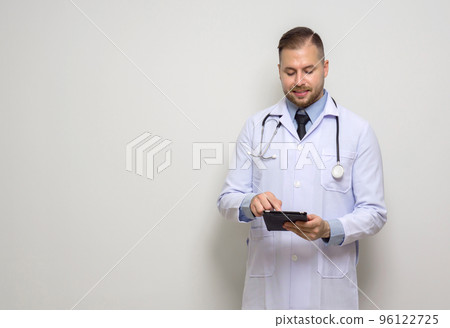 Young caucasian doctor with short hair, mustache and beard. Dressed  in white gown and stethoscope typing on tablet computer. Stand in front of white wall. 96122725