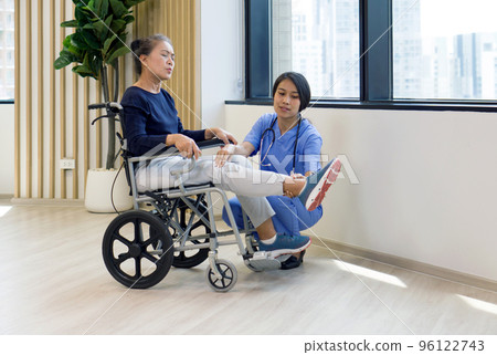 Young asian physical therapist in blue uniform help an elderly patient in a wheelchair lift her leg to relieve pain. 96122743