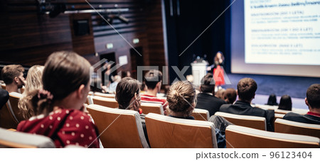 Business and entrepreneurship symposium. Female speaker giving a talk at business meeting. Audience in conference hall. Rear view of unrecognized participant in audience. 96123404