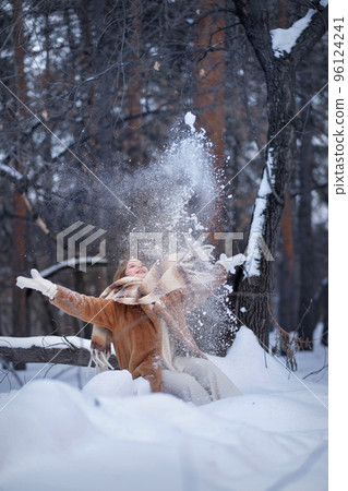 A white blonde girl in a knitted hat, a long scarf and a fur coat walks through the forest and smiles. Woman catches snow in white mittens. Winter is on the street. High quality photo. Christmas Eve 96124241