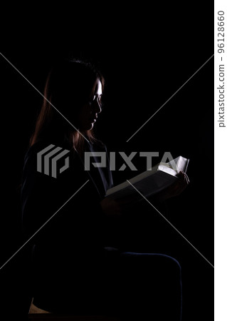 Vintage silhouette young beautiful woman sitting at night on chair and think about information reading. Glasses female read thick book bible and pray in the Dark. Black background isolated Vintage silhouette young beautiful woman sitting at night on chair and think about information reading. Glasses female read thick book bible and pray in the Dark. Black background isolated 96128660