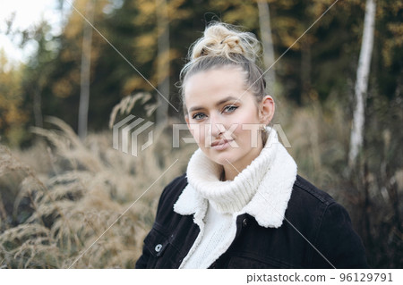 Lifestyle portrait of beautiful long hair blond caucasian girl, young woman in white knitted sweater, black jacket. Dry grass meadow. Autmn, fall landscape with yellow birch trees. Blurred woodland. Lifestyle portrait of beautiful long hair blond caucasian girl, young woman in white knitted sweater, black jacket. Dry grass meadow. Autmn, fall landscape with yellow birch trees. Blurred woodland. 96129791