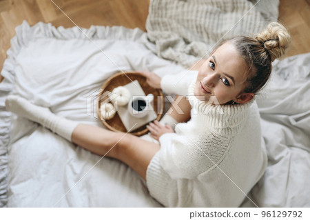 Beautiful blond caucasian girl, young woman in white knitted sweater, socks holding cup of coffee. Winter, autumn breakfast in bed. Wicker tray, white little pumpkins. Blurred background, top view. Beautiful blond caucasian girl, young woman in white knitted sweater, socks holding cup of coffee. Winter, autumn breakfast in bed. Wicker tray, white little pumpkins. Blurred background, top view. 96129792