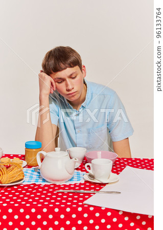 Portrait of young boy in blue shirt sitting at the table and having breakfast isolated on grey background. Bored 96133764