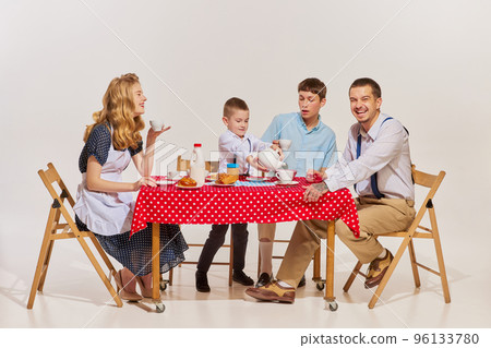 Portrait of lovely young family, woman, man and two boys sitting at the table and having breakfast isolated over grey background. Morning talk 96133780