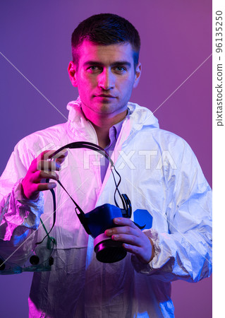 Coronavirus covid-19 pandemic. Doctor scientist wearing protective biological suit and mask due to global healthcare epidemic warning and danger background in blue and pink neon lights background. Coronavirus covid-19 pandemic. Doctor scientist wearing protective biological suit and mask due to global healthcare epidemic warning and danger background in blue and pink neon lights background. 96135250