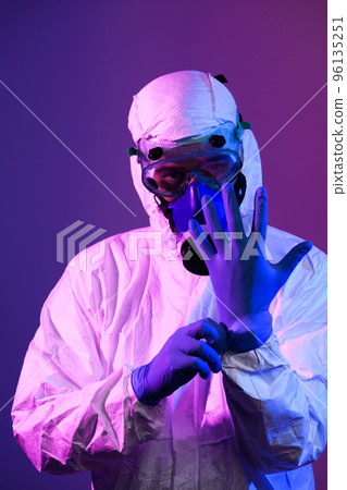 Coronavirus covid-19 pandemic. Doctor scientist wearing protective biological suit and mask due to global healthcare epidemic warning and danger background in blue and pink neon lights background. Coronavirus covid-19 pandemic. Doctor scientist wearing protective biological suit and mask due to global healthcare epidemic warning and danger background in blue and pink neon lights background. 96135251