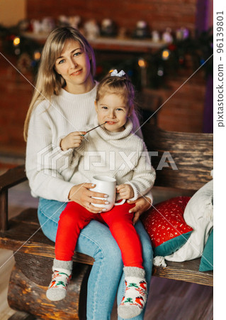 Portrait of a happy little girl sitting on her mother's lap and drinking hot chocolate on Christmas or New Year's Eve. Christmas mood, Christmas spirit, time together, motherhood, childhood 96139801